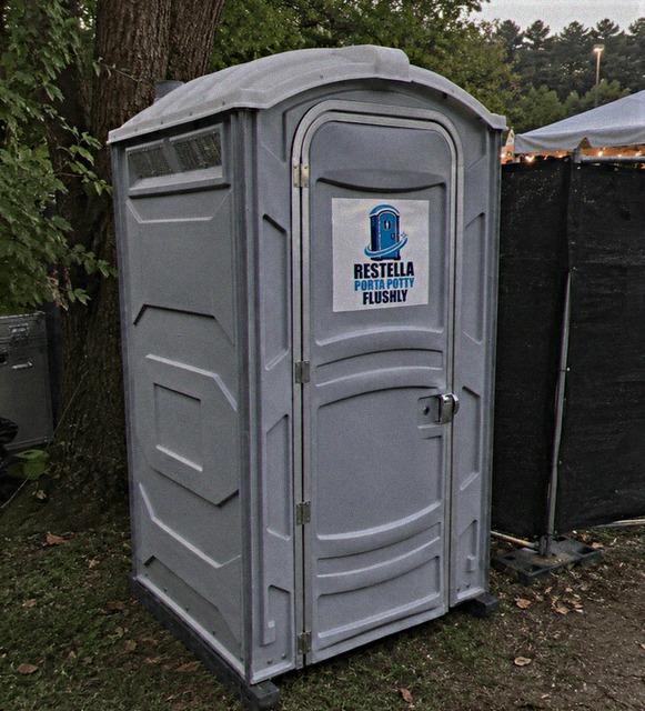 Grey portable restroom placed on grass beside a marquee at an outdoor gathering